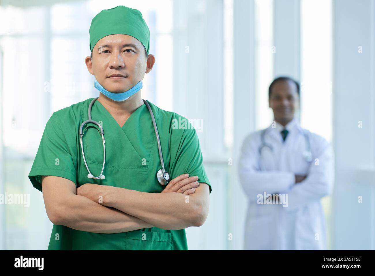 Médecin sérieux dans des maquillages verts croise les bras et regarde la caméra. Ce portrait professionnel exprime l'expertise et la confiance dans le domaine de la santé, idéal pour les cliniques, les hôpitaux et le marketing médical. Adapté aux campagnes médicales d'entreprise, aux profils du personnel et aux communications centrées sur le patient. Banque D'Images