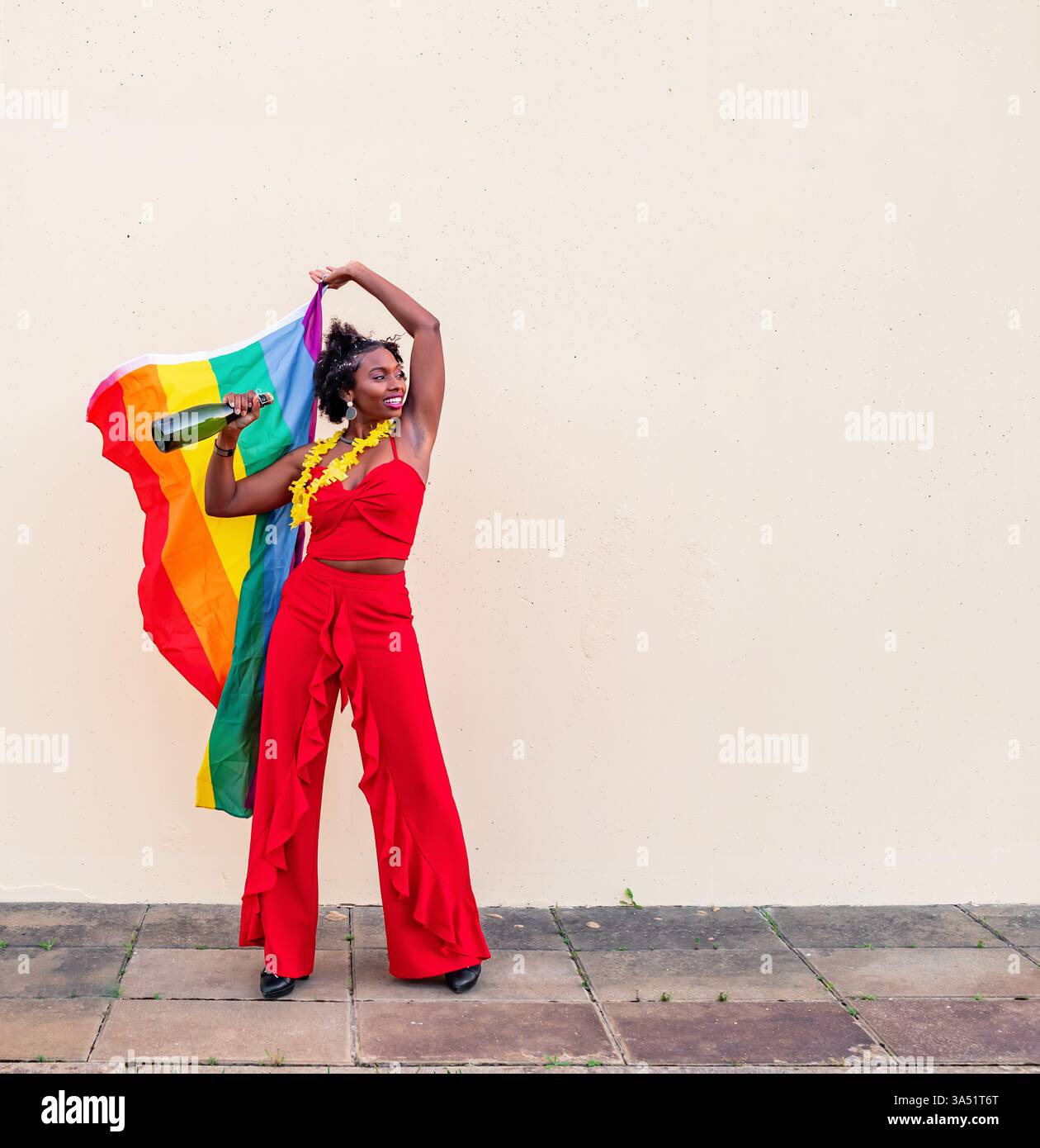 Femme noire souriante célébrant la fierté avec un drapeau LGBTQ et une boisson contre un mur blanc. Une image colorée et inclusive de style de vie idéale pour les campagnes de fierté, la sensibilisation LGBTQ et la narration de marque. Idéal pour les médias sociaux, les événements et les messages d'égalité. Banque D'Images