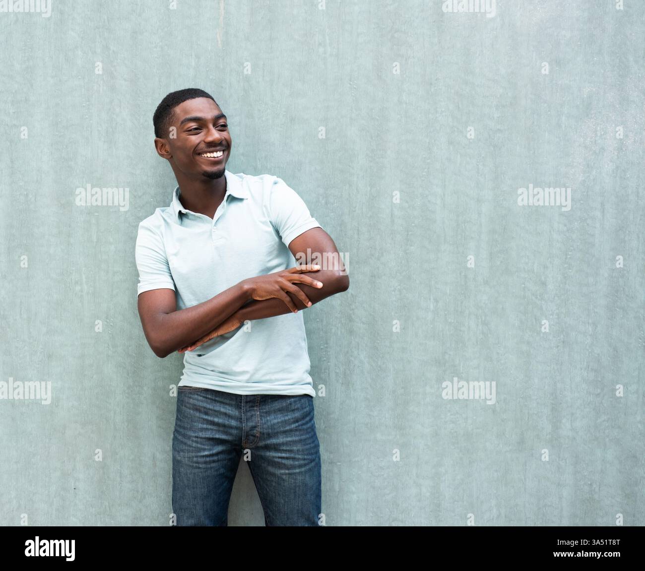 Homme noir souriant avec les bras croisés debout contre un mur gris, regardant loin. Un portrait de style de vie contemporain avec une ambiance confiante et détendue adapté à la mode, la publicité et la marque personnelle. Banque D'Images