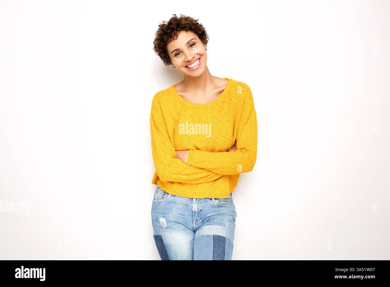 Femme afro-américaine souriante avec les bras croisés contre un mur blanc. Ce portrait de style de vie décontracté transmet confiance et style pour la marque personnelle ou le contenu de mode. Banque D'Images