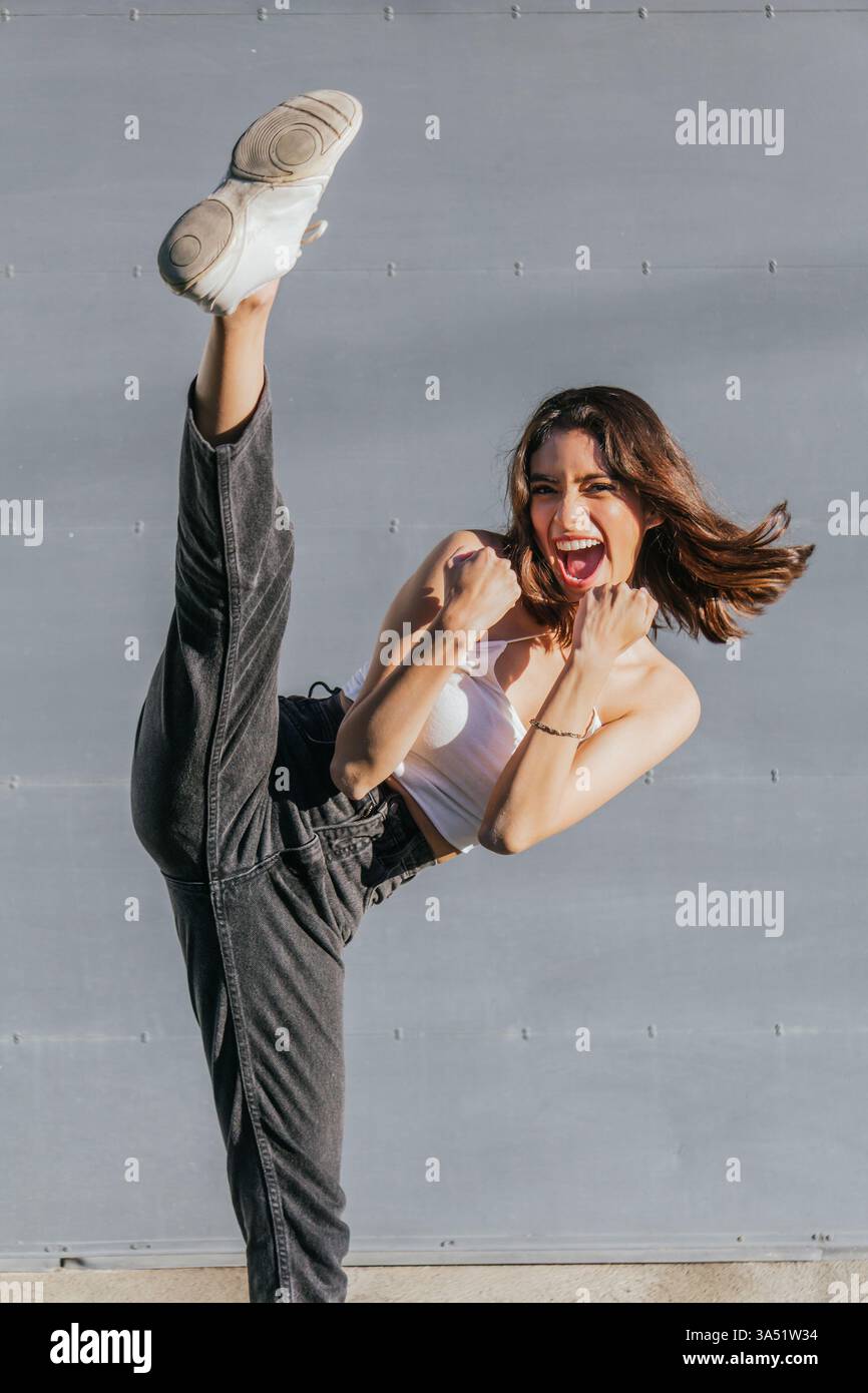 Image énergique en plein air d'une femme hispanique en tenue de fitness effectuant une pose dynamique avec une jambe levée et les poings serrés. Idéal pour les campagnes sportives, de fitness et de style de vie actif, véhiculant énergie et motivation en extérieur. Banque D'Images