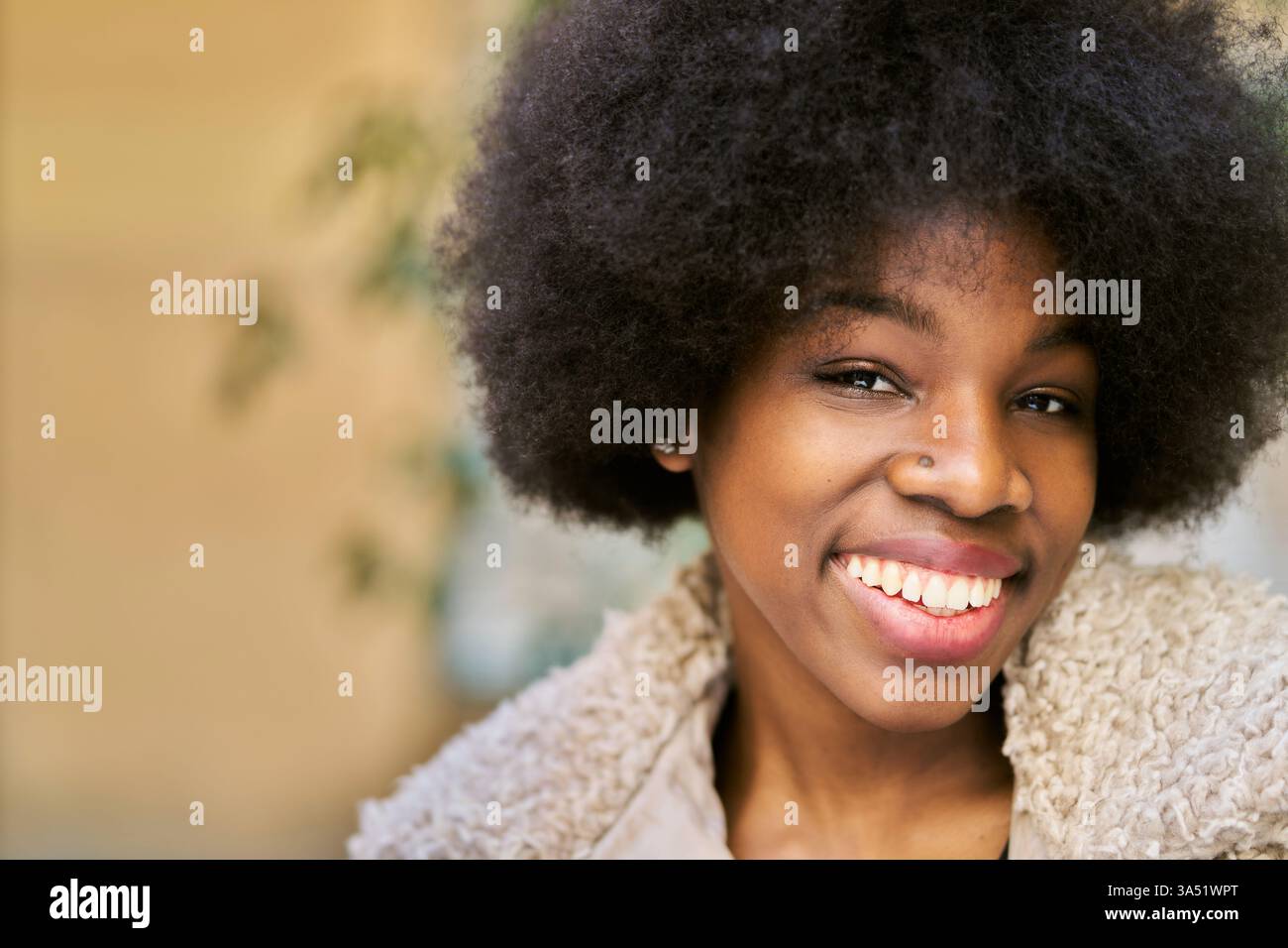 Joyeuse femme afro-américaine avec une coiffure afro sourit à la caméra dans une tenue confortable et décontractée. Le portrait est photographié en extérieur avec un arrière-plan flou et doux, ce qui le rend idéal pour un style de vie, une beauté ou une utilisation de marque personnelle. Un coup de tête chaleureux qui communique la convivialité et la confiance. Banque D'Images