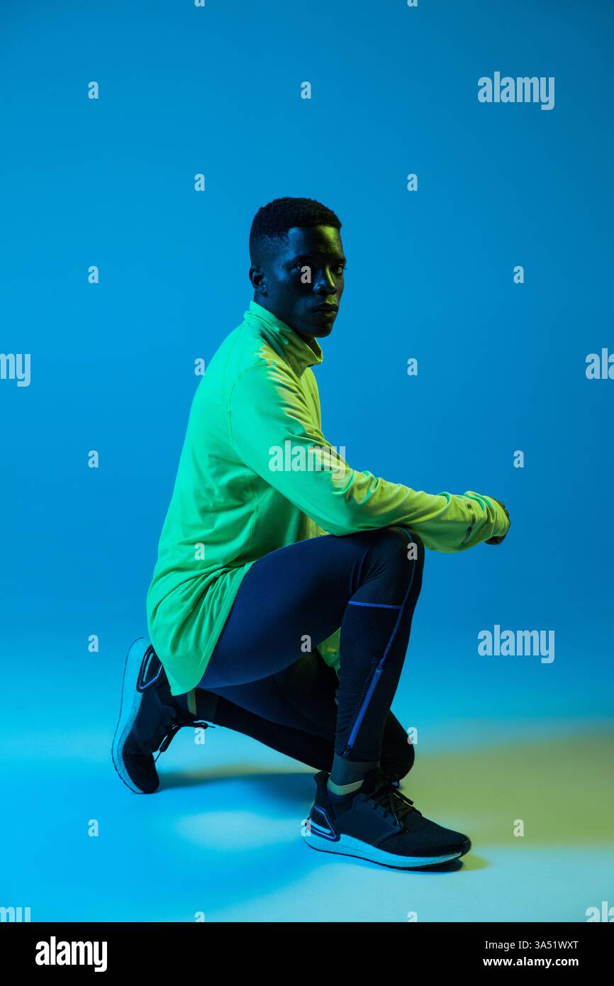 Jeune homme noir dans un survêtement néon debout sur un genou dans un studio. La pose athlétique met l'accent sur le fitness, les vêtements de sport et l'entraînement. Idéal pour les campagnes sportives et les images de style de vie actif. Banque D'Images