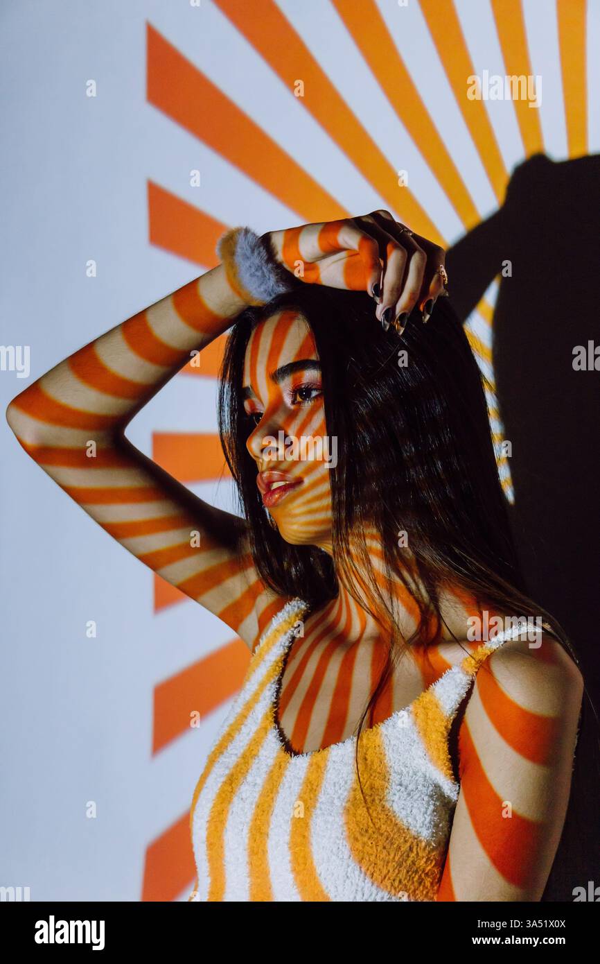 Femme hispanique avec la main dans les cheveux contre une rayure reflétant un mur. Ce portrait de style de vie communique confiance et individualité, adapté à la mode et au contenu de style personnel. Banque D'Images
