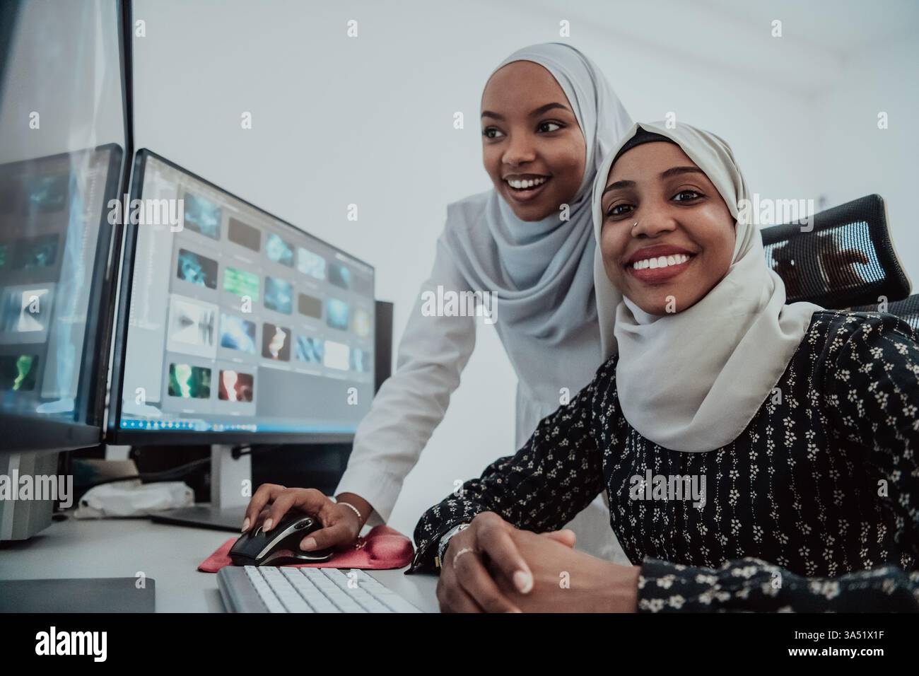 Deux jeunes femmes d'affaires afro-américaines portant des foulards travaillent dans un bureau lumineux et créatif avec un grand écran. Ce milieu de travail moderne convient au travail d'équipe, aux femmes dans le leadership, à la diversité et aux campagnes de communication d'entreprise. Banque D'Images