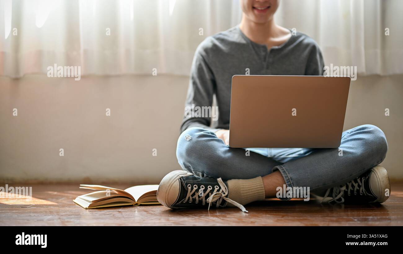 Homme asiatique travaillant sur un ordinateur portable, assis les jambes croisées sur le sol à la maison pendant la journée. Scène de bureau à domicile décontracté adaptée aux contextes de travail et d'étude à distance. Idéal pour les freelances, l'apprentissage en ligne et les campagnes de vie confortable. Banque D'Images