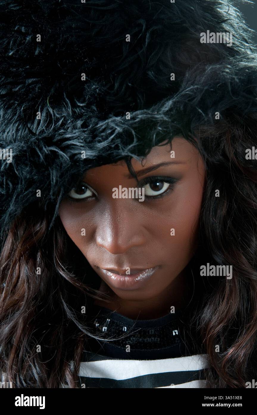 Femme noire sérieuse portant un chapeau noir se tient sur un fond noir solide. Un portrait audacieux et minimaliste adapté à la mode, à la beauté ou aux campagnes éditoriales. Idéal pour des visuels d'identité forts et des images de style dramatique. Banque D'Images