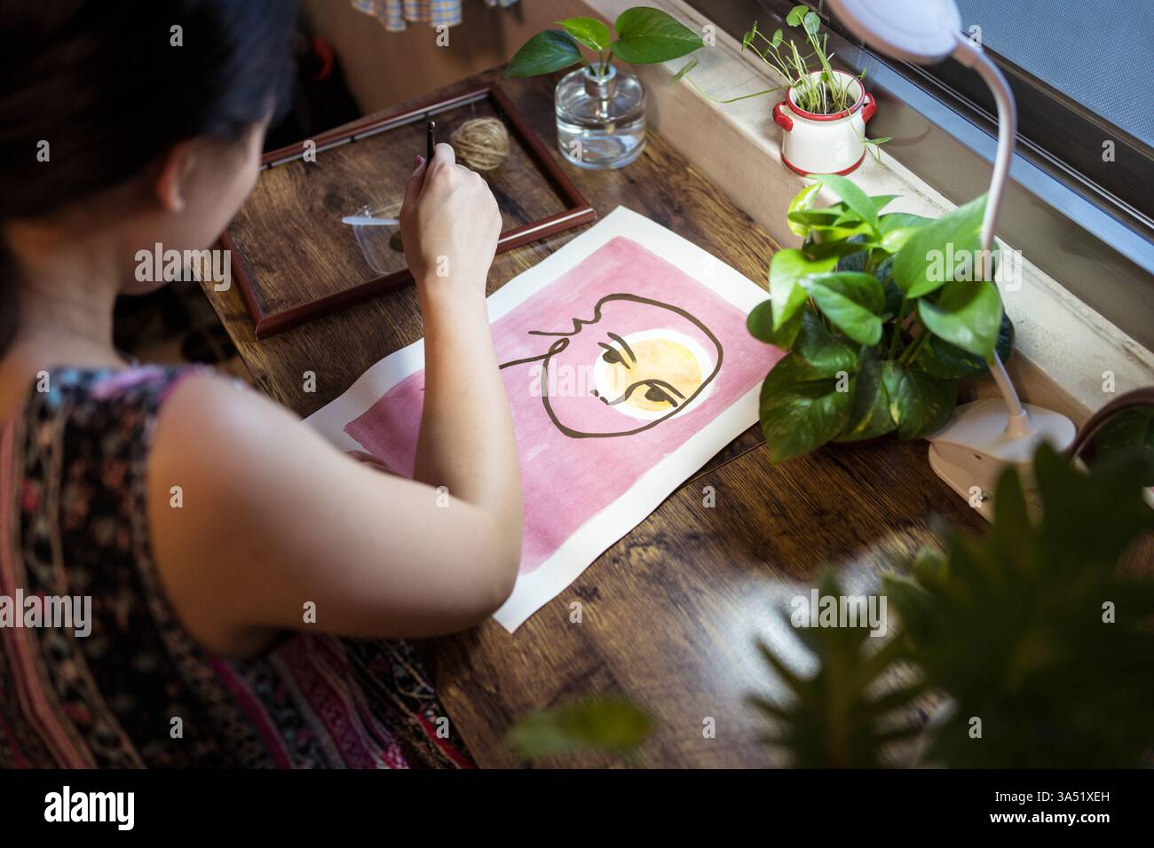 Artiste femme asiatique assise à une table en bois et peignant sur papier avec un pinceau. Cette image de style studio met en valeur la créativité, l'artisanat et les œuvres d'art faites à la main. Idéal pour les fonctionnalités sur l'art, les techniques de peinture et les home studios. Banque D'Images