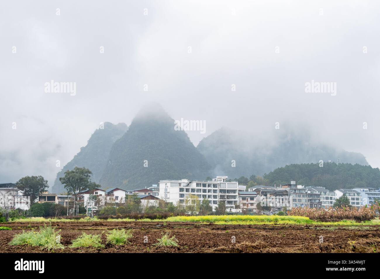 Les montagnes brumeuses et les champs dynamiques encadrent un village chinois serein, capturant l'harmonie de la nature et de la vie rurale. Banque D'Images Les montagnes brumeuses et les champs dynamiques encadrent un village chinois serein, capturant l'harmonie de la nature et de la vie rurale. Banque D'Images