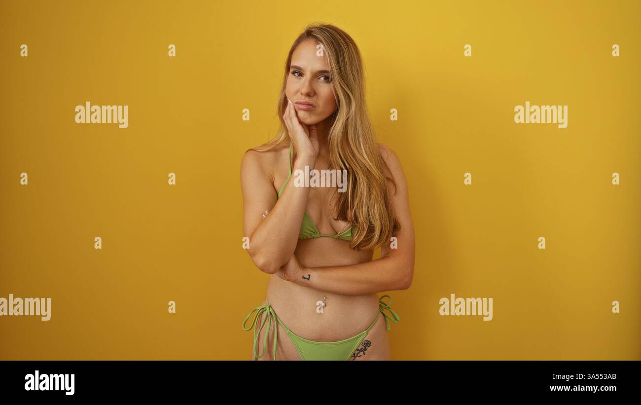 Belle jeune femme aux longs cheveux blonds dans un bikini vert posant contre un mur jaune vif, véhiculant une expression réfléchie et un ensemble isolé Banque D'Images