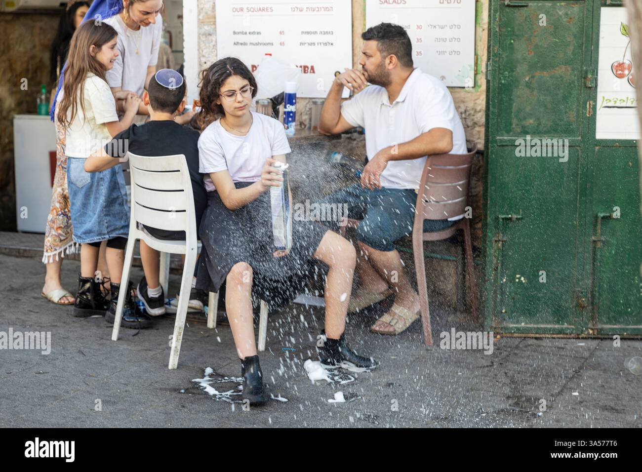 Jérusalem, Israël – 16 mars 2025. Une grande famille dans un café. Pendant que les parents choisissent de la nourriture, une fille éclabousse de la mousse neigeuse. Banque D'Images