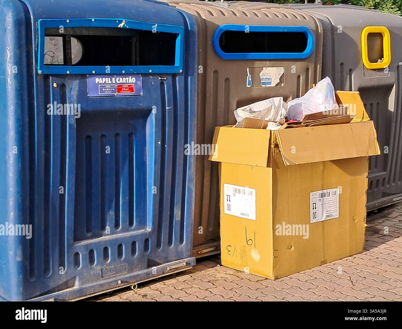 La boîte en carton débordante remplie de déchets de papier et de plastique se trouve à côté des bacs de recyclage à code couleur, soulignant les défis de gestion des déchets Banque D'Images