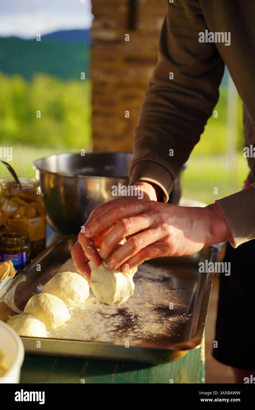 Les mains façonnent soigneusement des boules de pâte sur un plateau fariné, avec des ingrédients de cuisine tels que des pots et un bol à mélanger à proximité, sur un dos extérieur pittoresque Banque D'Images