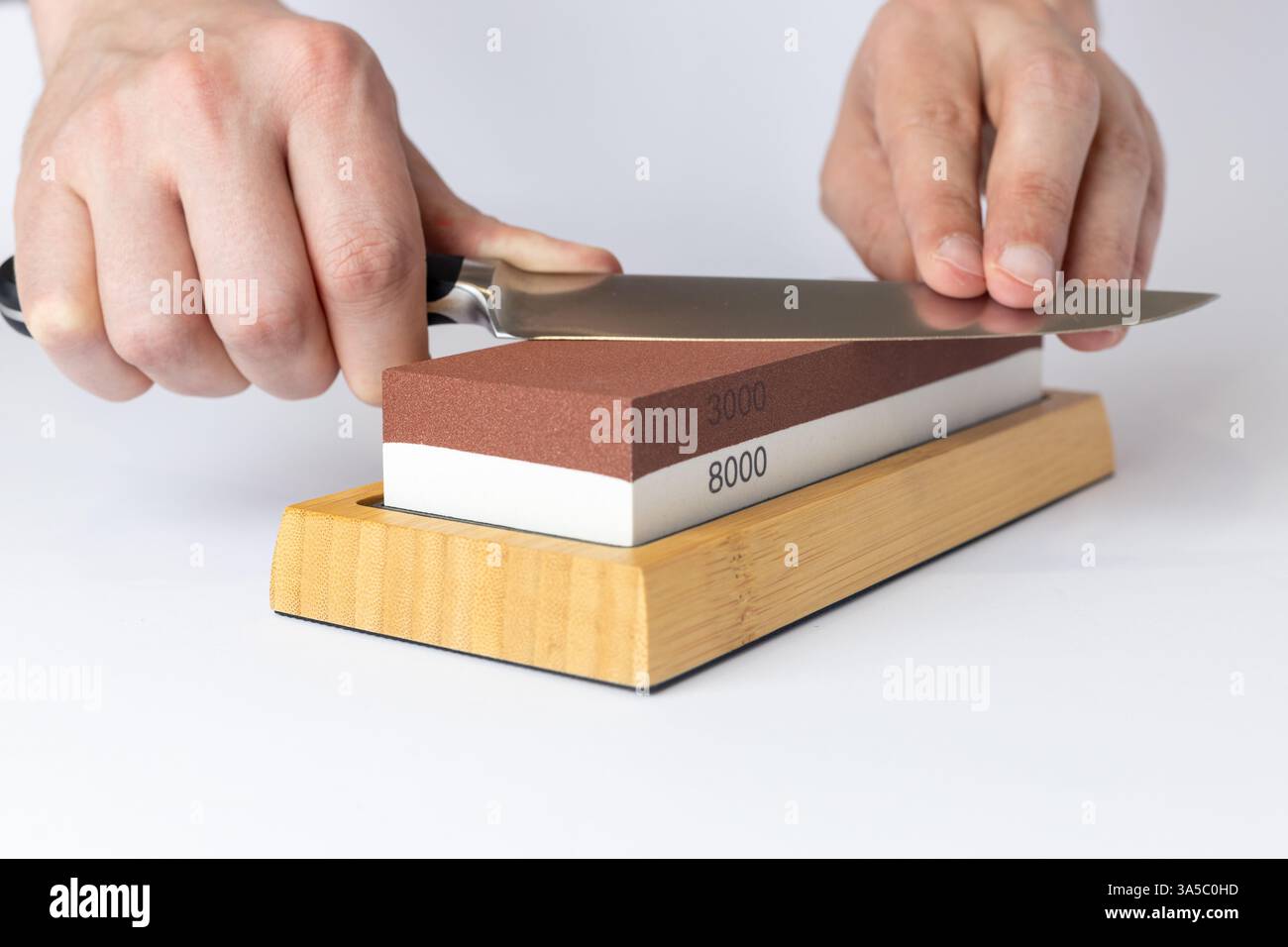 gros plan sur fond blanc sur une table, les mains de l'homme affûtent une protection de couteau en acier contre les coupures, une pierre rouge moyennement dure pour affûter les couteaux Banque D'Images