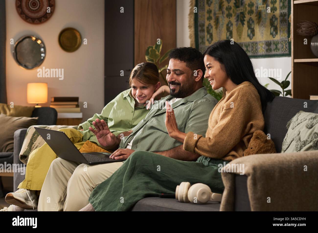 Groupe de trois personnes assis sur le canapé, engagé avec un ordinateur portable, souriant et agitant à l'écran dans un environnement de salon confortable décoré avec un éclairage chaleureux Banque D'Images