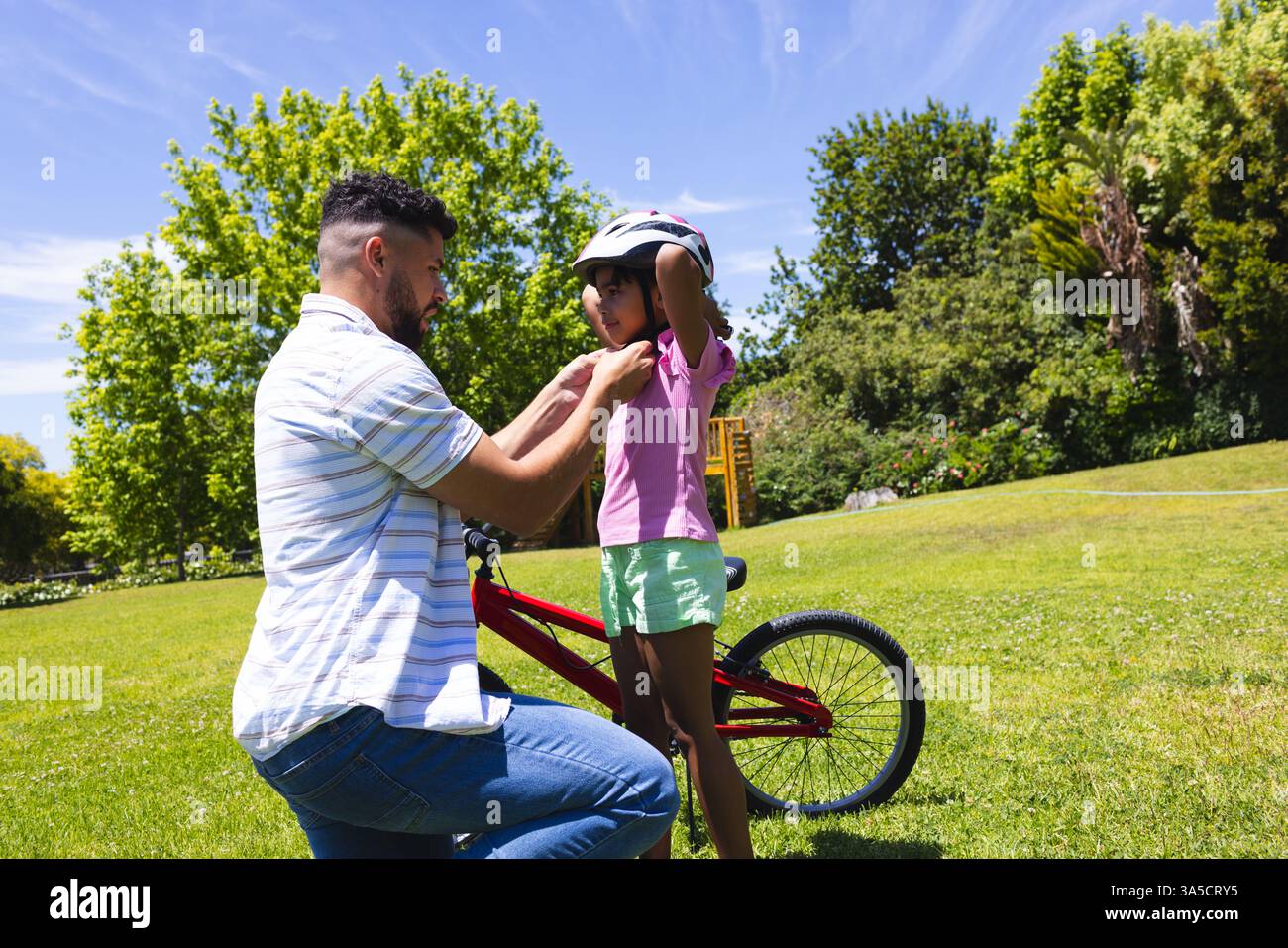 Père aidant la fille avec casque avant la balade à vélo dans le parc ensoleillé Banque D'Images