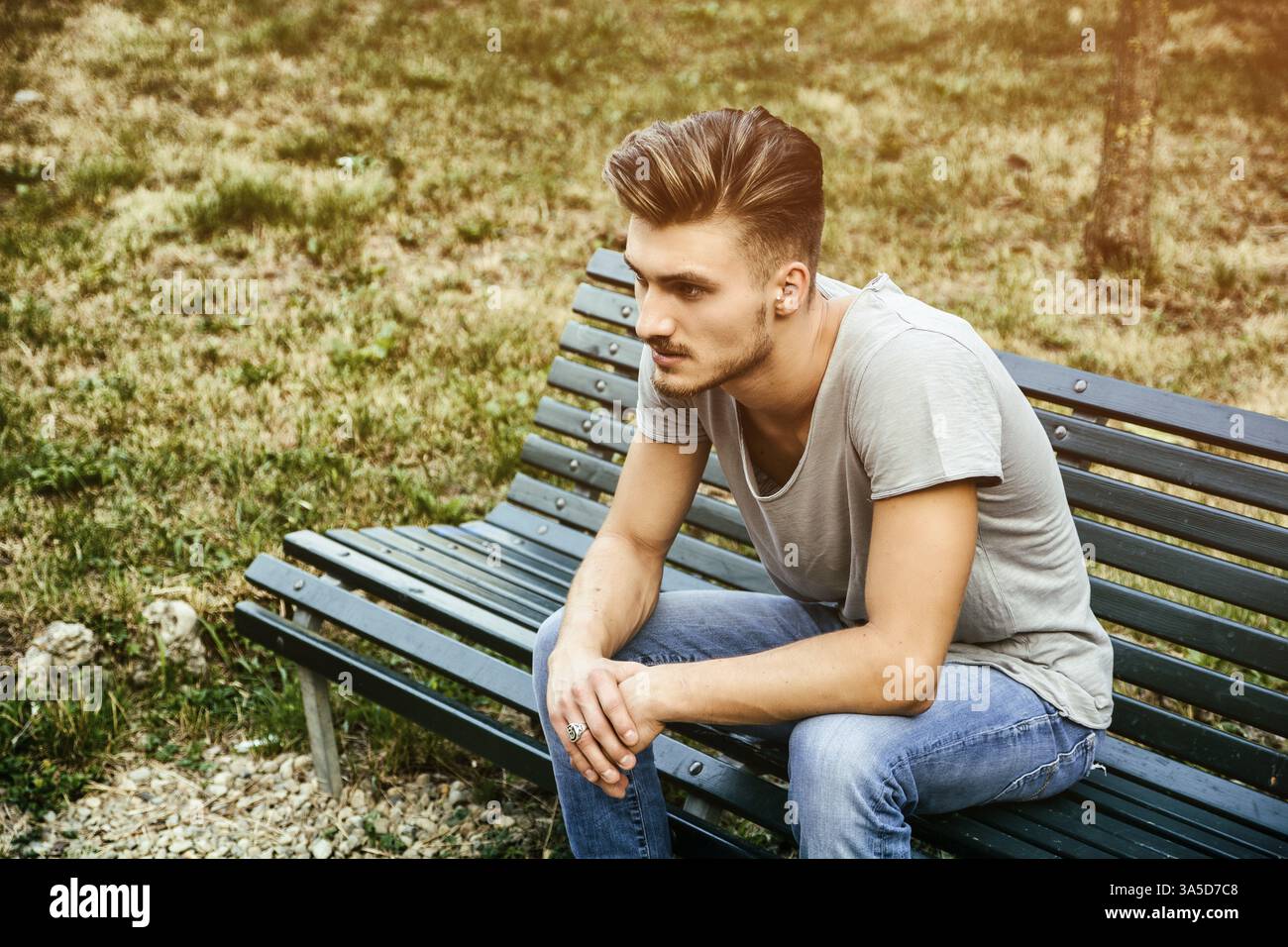 Beau jeune homme blond assis sur le banc de parc en bois, vert Banque D'Images