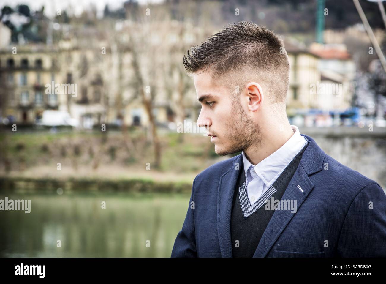 Trois quarts de longueur de jeune homme contemplatif à poil brun clair portant une veste grise et un Jean denim debout à côté de la rivière pittoresque à Turin, ITA Banque D'Images