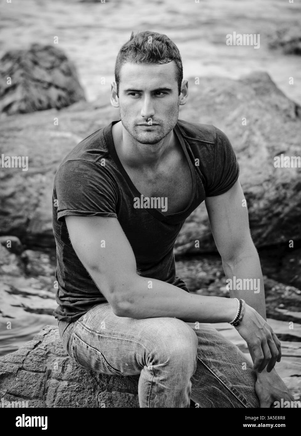 Portrait en noir et blanc d'un jeune homme attrayant et athlétique assis sur un rocher au bord de la mer Banque D'Images