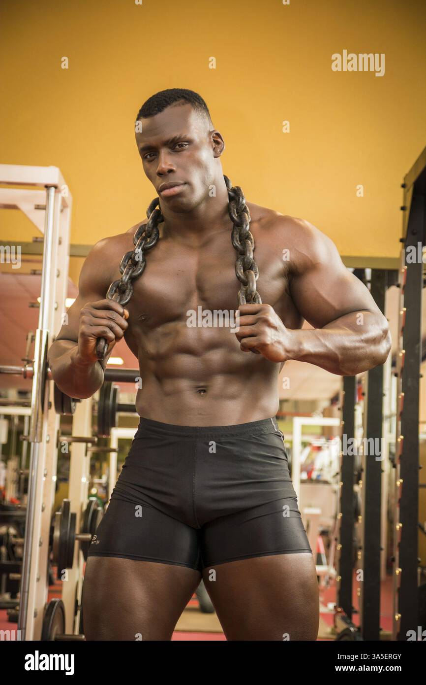Belle black hunky male bodybuilder bodybuilding faire poser dans une salle de sport avec des chaînes de fer sur les épaules Banque D'Images