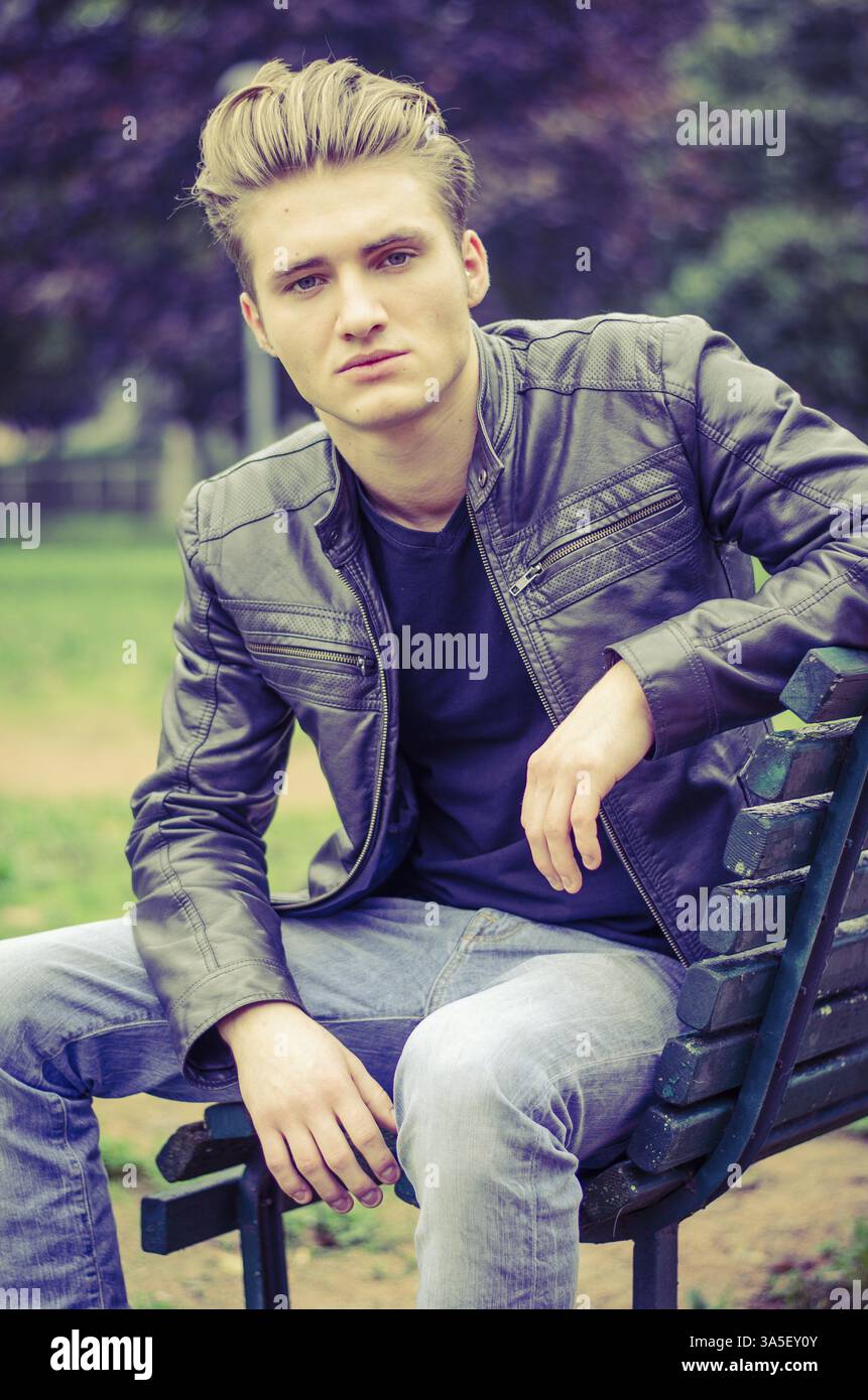 Beau jeune homme blond assis sur le banc de parc en bois, vert Banque D'Images