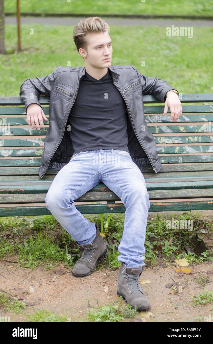 Beau jeune homme blond assis sur un banc vert de parc en bois regardant loin Banque D'Images