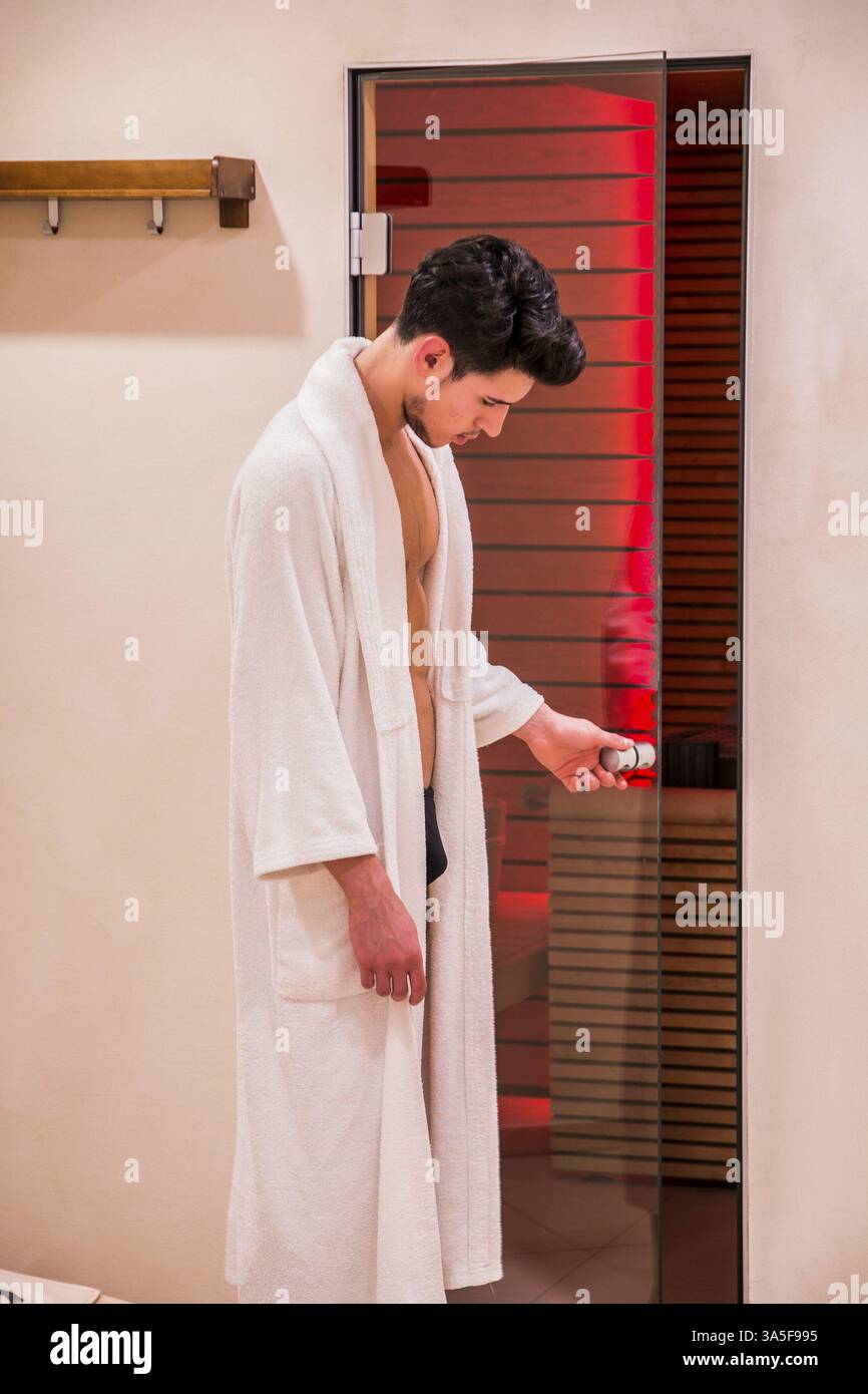 Beau jeune homme en blanc peignoir entrant ou sortant d'un sauna, l'ouverture d'une porte en verre pour entrer ou sortir Banque D'Images