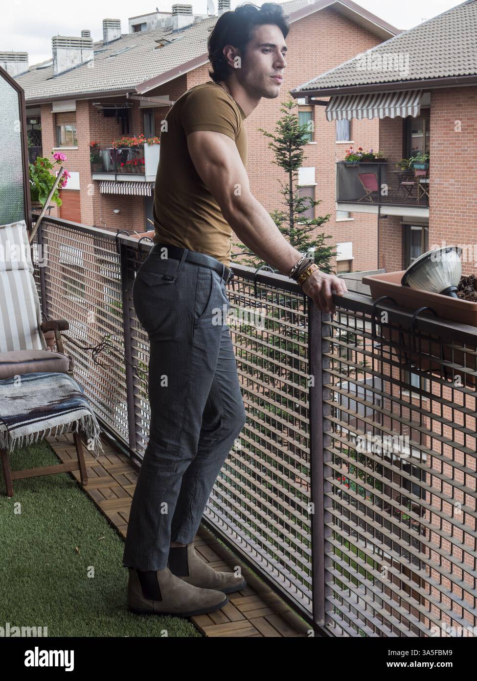 Beau jeune homme aux cheveux foncés debout dans un balcon, regardant dehors et en bas à la vue, portant un T-shirt marron, en été, plan en longueur Banque D'Images