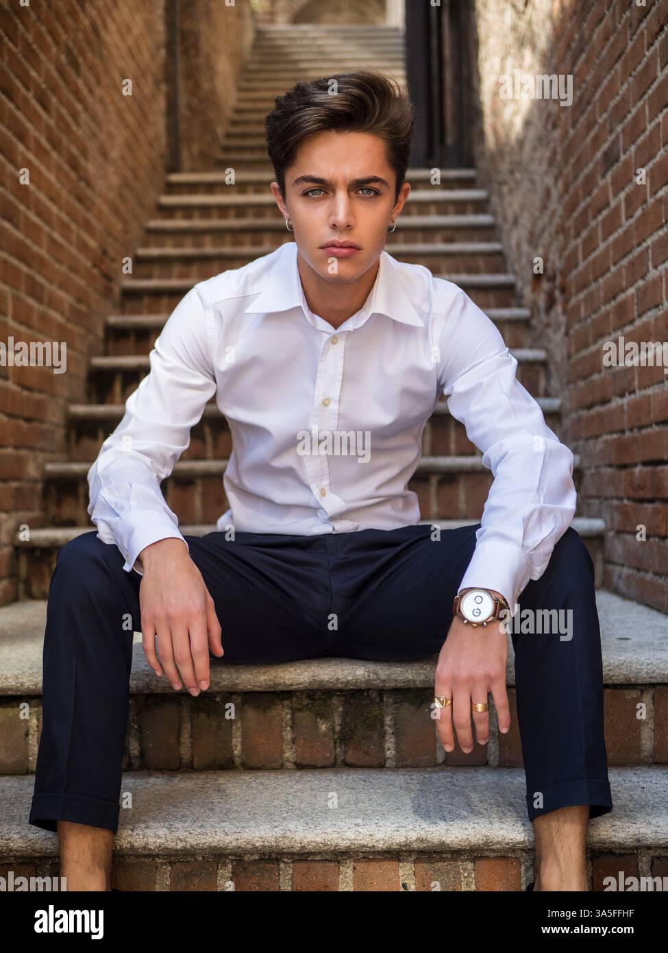 Un beau jeune homme élégant dans un cadre urbain dans la ville européenne, assis sur de vieux escaliers, regardant la caméra Banque D'Images