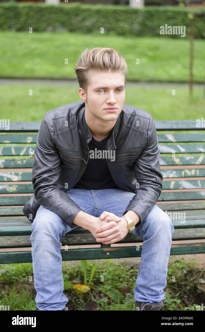 Beau jeune homme blond assis sur le banc de parc en bois, vert Banque D'Images