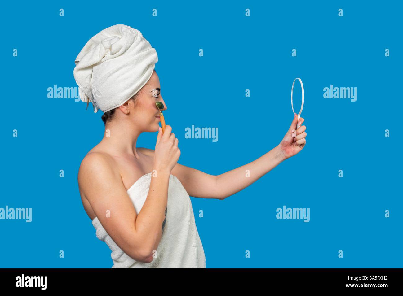 Profil de femme souriante enveloppée dans une serviette à l'aide d'un rouleau de jade et d'un miroir, profitant de sa routine de beauté après la douche Banque D'Images