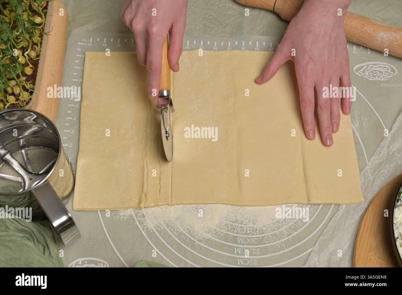 Les mains de la femme coupent une feuille de pâte. Rouleau à pâtisserie, couteau, serviette verte, germes de microgreens sur la table de cuisine. Maison cuisson pâtisseries feuilletées Banque D'Images