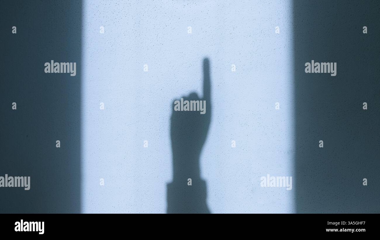 Ombre d'une main pointant vers le haut projetée par la lumière du soleil sur un mur, créant un contraste visuel saisissant avec un éclairage naturel et une composition minimaliste Banque D'Images