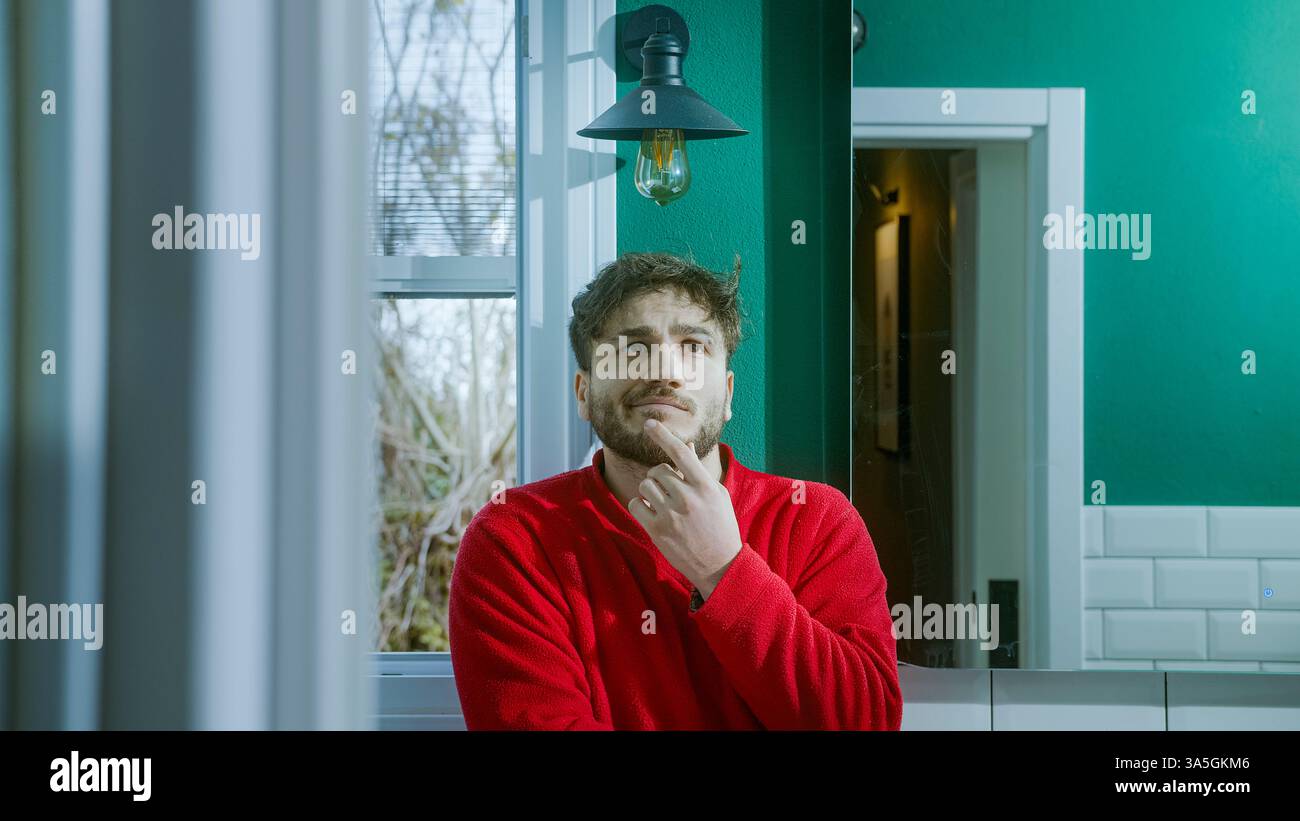 Jeune homme réfléchi dans un pull rouge debout dans une salle de bains élégante avec des murs verts, profond dans la contemplation. Banque D'Images