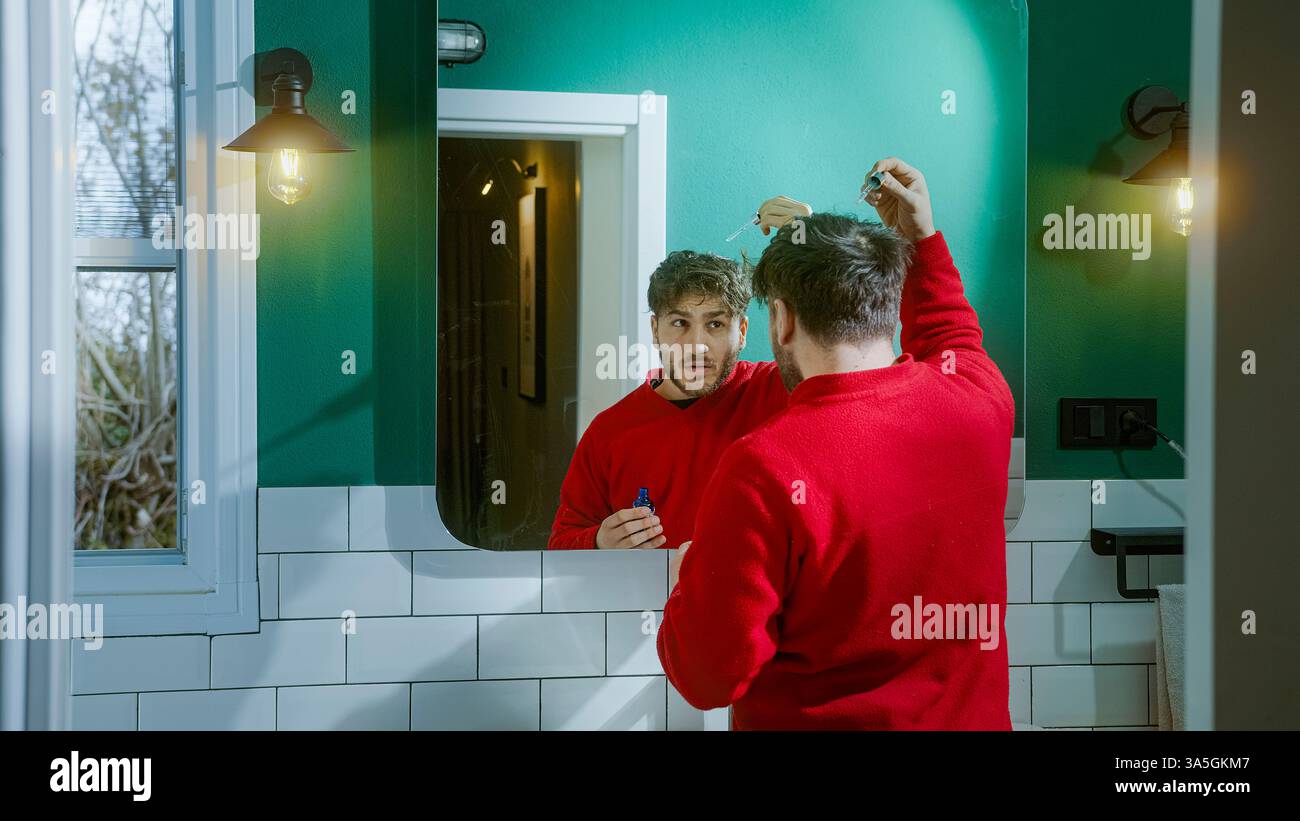 Homme dans un pull rouge appliquant soigneusement le sérum de soins capillaires devant un miroir, suivant une routine quotidienne de toilettage pour combattre la perte de cheveux et améliorer le cuir chevelu Banque D'Images