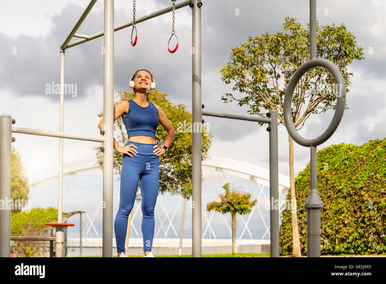 Jeune femme sportive dans des écouteurs regardant vers les anneaux de gymnastique dans un parc de fitness en plein air et souriant Banque D'Images