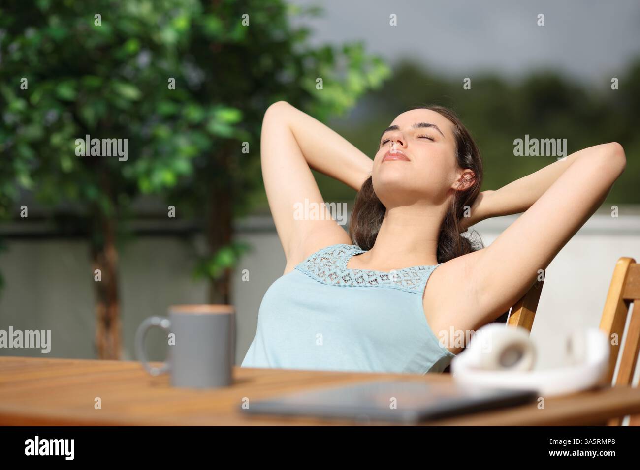 Femme se relaxant avec les bras sur la tête assis dans une terrasse dans une maison une journée ensoleillée Banque D'Images