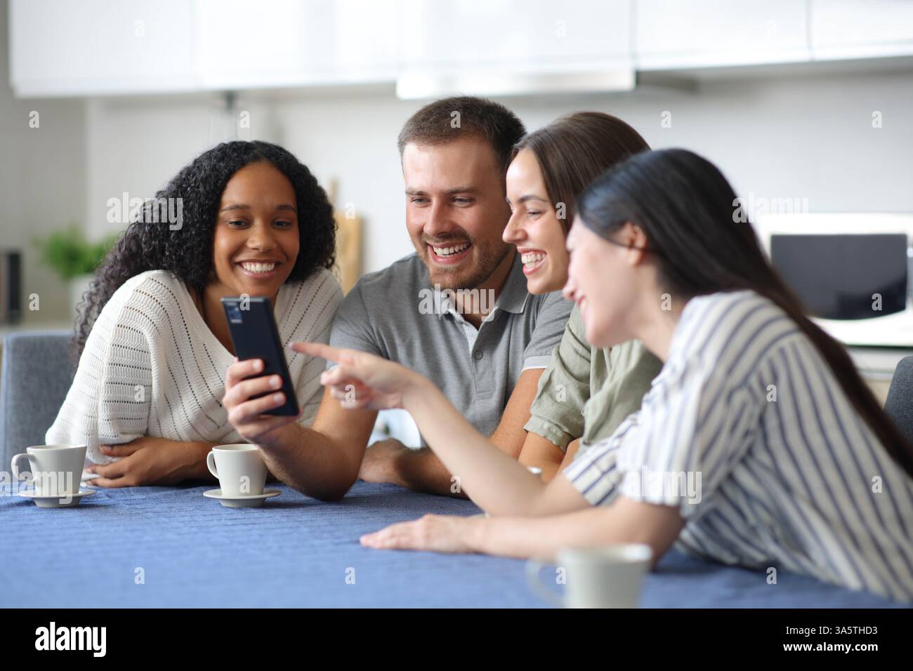 Quatre amis interraciaux heureux parlant du contenu du téléphone dans la cuisine à la maison Banque D'Images