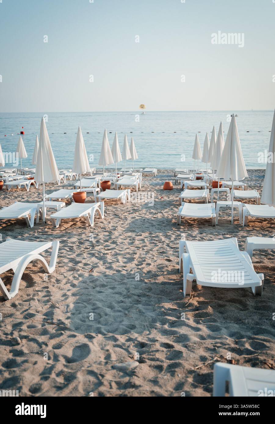 Une scène de plage tôt le matin avec des chaises longues vides et des parasols fermés disposés soigneusement sur le sable. La mer calme et le parasailer lointain ajoutent à la Banque D'Images