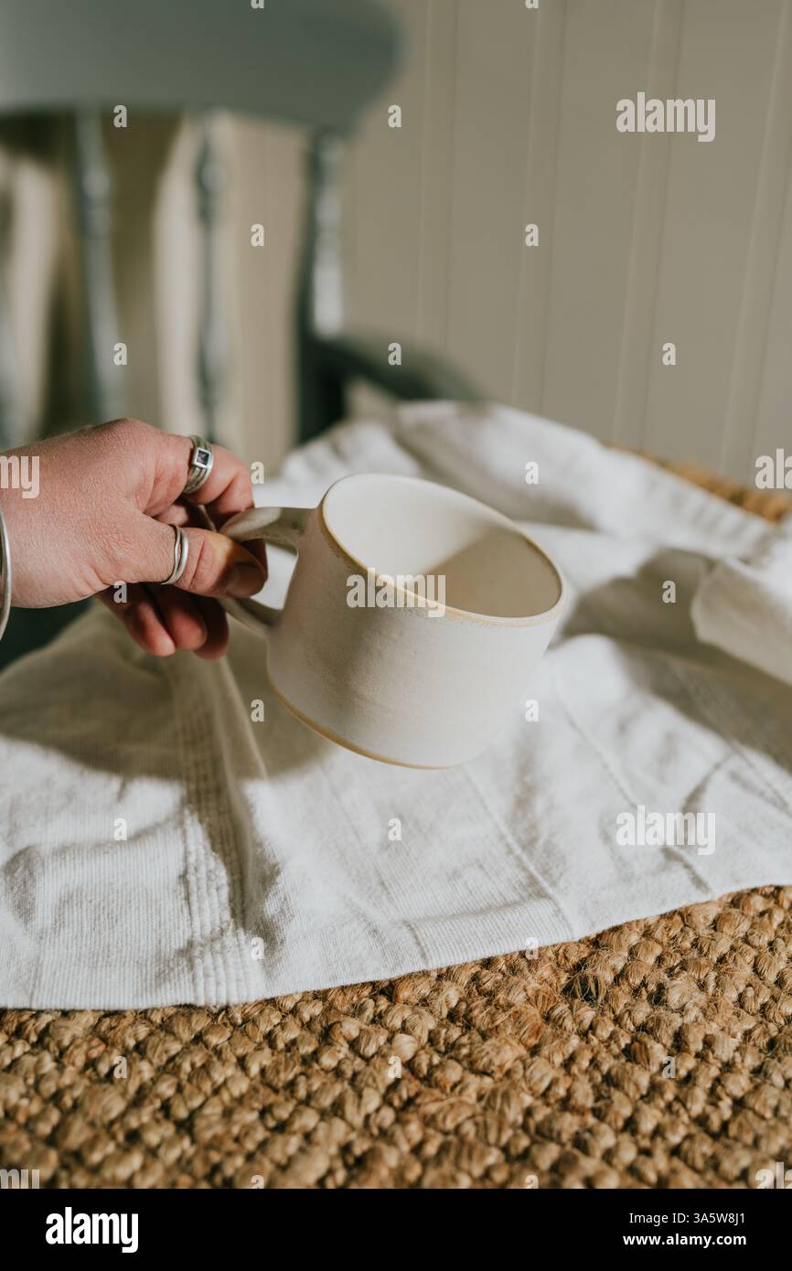 Une main tenant une tasse blanche sur un chiffon blanc. La tasse est placée sur un tapis. La tasse est blanche et a un bord jaune Banque D'Images