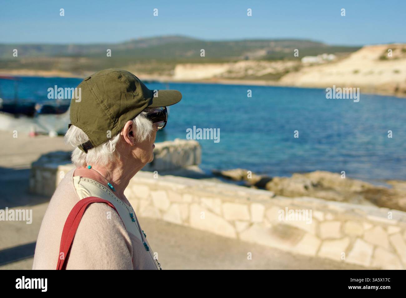 Femme âgée profitant d'une vue sereine sur la côte, portant un chapeau vert et une tenue décontractée sous le ciel bleu vif, exsudant le calme et l'appréciation Banque D'Images