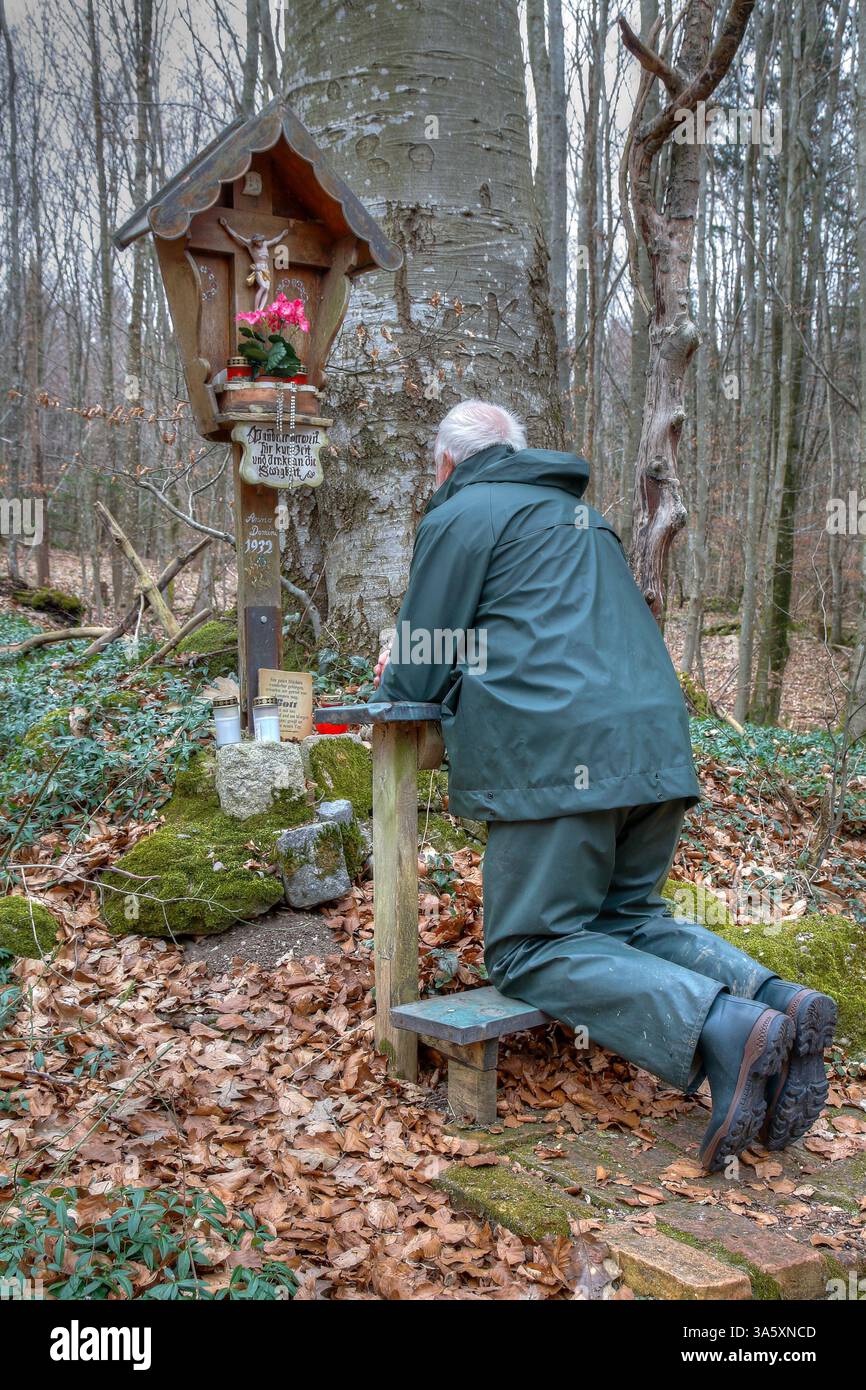 Un randonneur s'agenouille devant une croix en bois dans la forêt bavaroise. Vêtu de vêtements de pluie, il reste dans la prière silencieuse. Banque D'Images