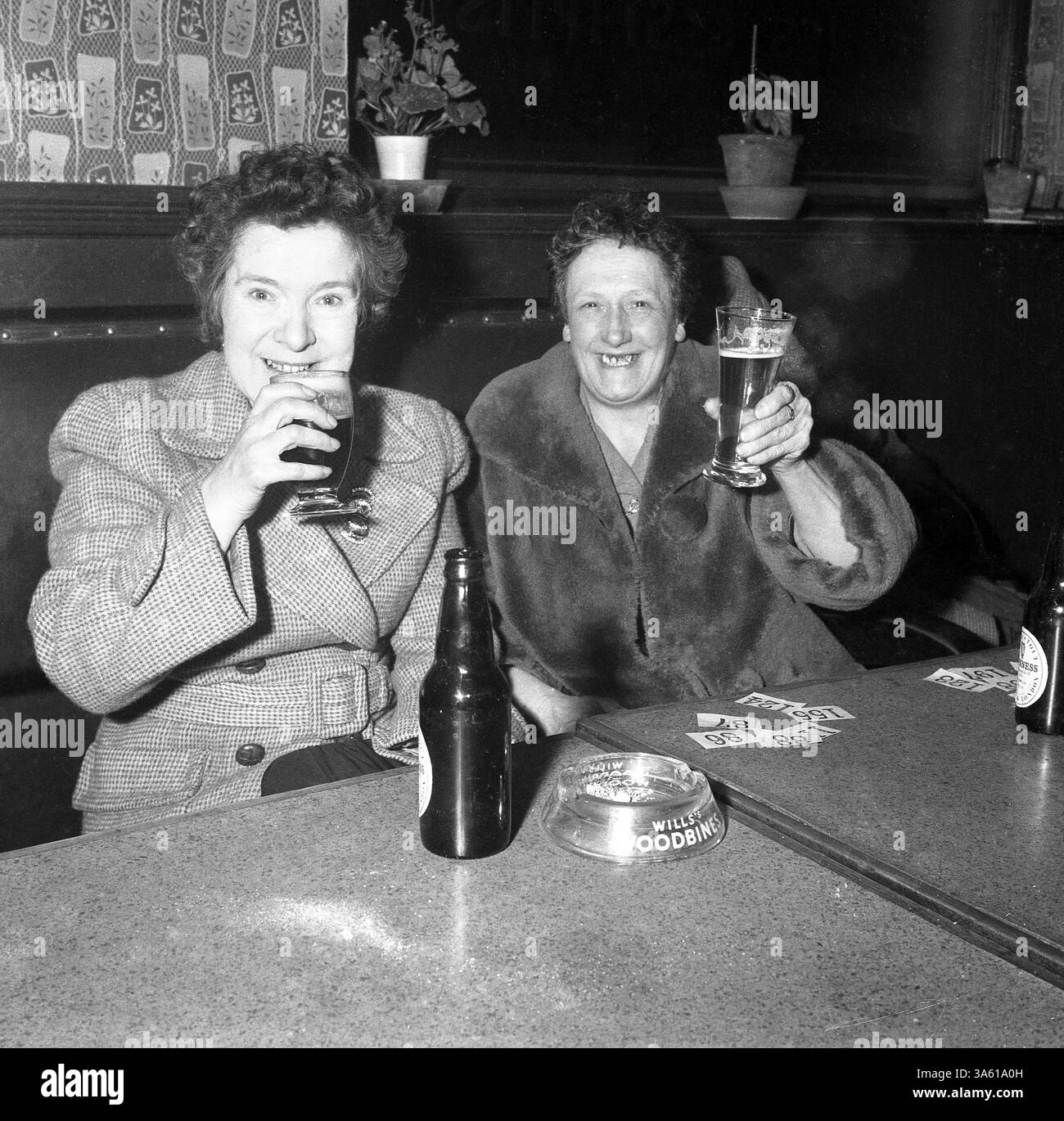 années 1950, historique, acclamations.... Deux dames, toutes deux portant un manteau, dont une en fourrure, lèvent leurs lunettes tout en dégustant un verre dans un pub East End London, Angleterre, Royaume-Uni. Bouteilles de bière, cendrier en verre et billets de tombola sur les tables. Banque D'Images