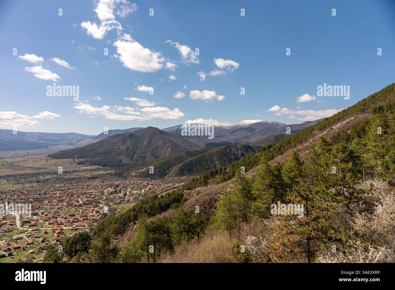 Beau paysage montrant une chaîne de montagnes avec des sommets enneigés et un village niché dans la vallée ci-dessous, sous un ciel bleu avec clou blanc moelleux Banque D'Images