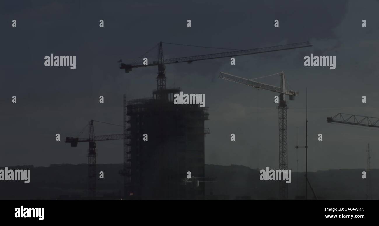 Grues de construction surplombant un immeuble de grande hauteur sous un ciel sombre et nuageux Banque D'Images