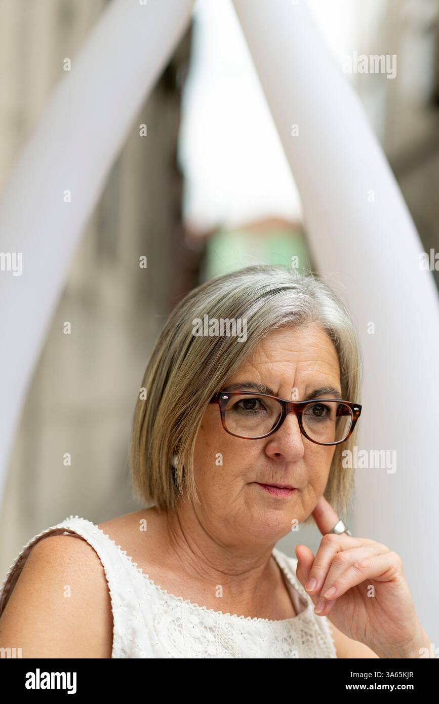 Une femme âgée avec des lunettes et des cheveux gris courts est assise à l'extérieur, profondément dans la pensée. Elle respire la sagesse et l'introspection dans un cadre urbain franc, fatigué Banque D'Images