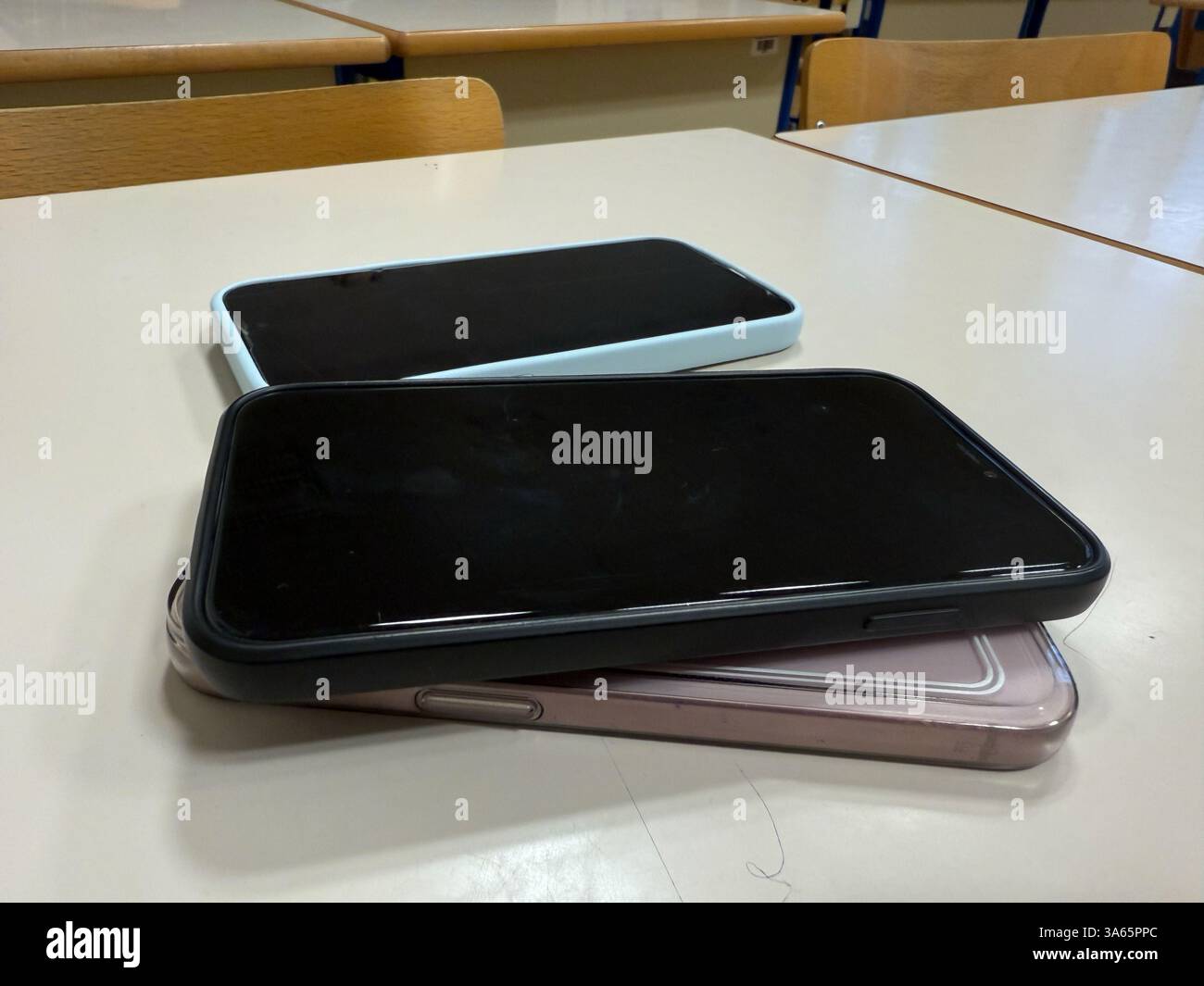 Gros plan de trois smartphones empilés sur un bureau dans une salle de classe vide, symbolisant une politique d'interdiction de téléphone ou une règle de collecte d'appareils pendant les heures d'école. Banque D'Images