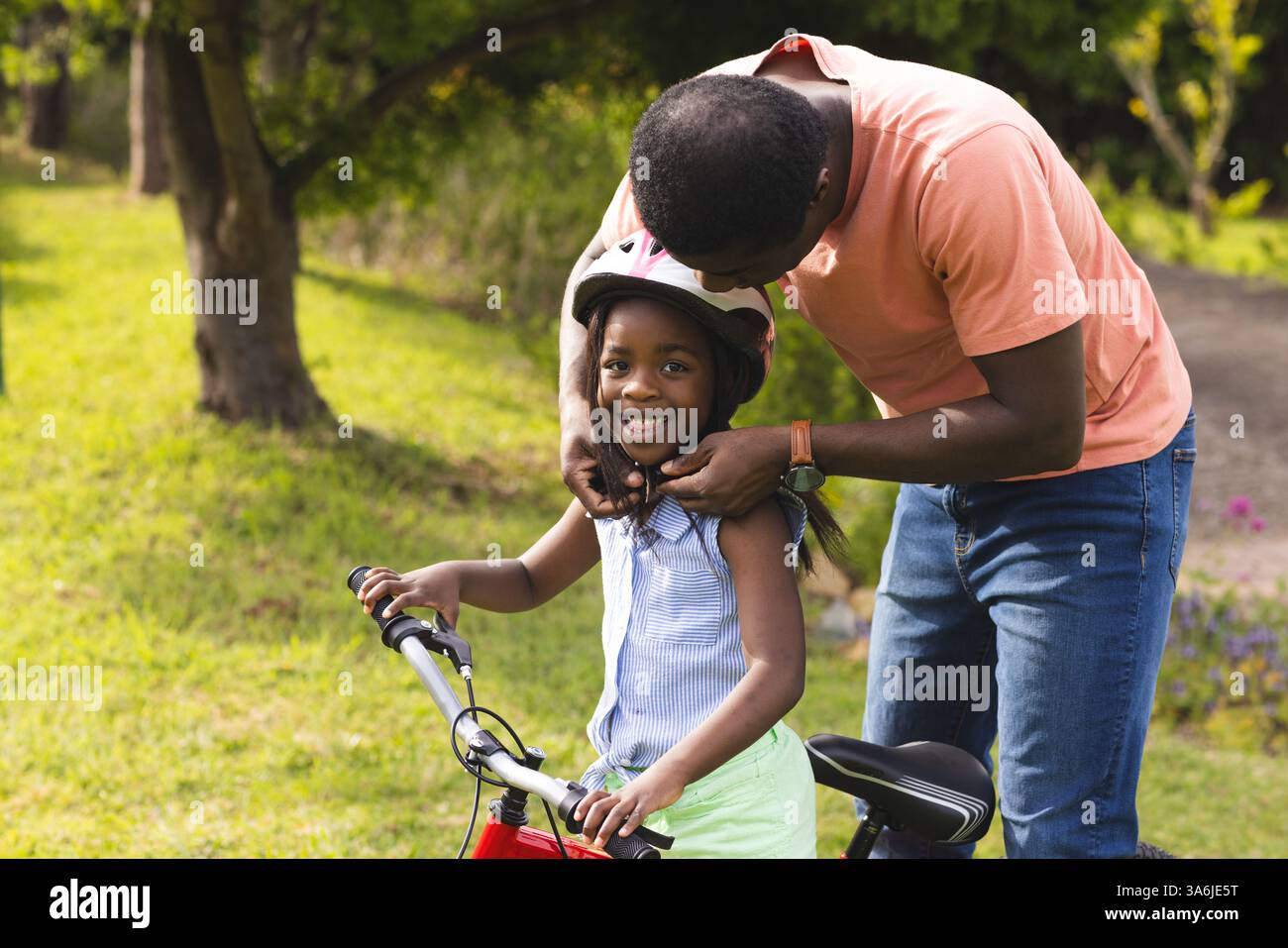 Père aidant sa fille avec un casque alors qu'elle sourit sur le vélo à l'extérieur Banque D'Images
