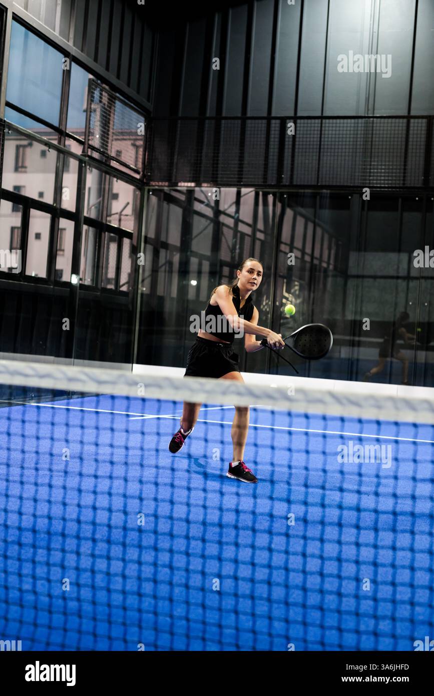 Femme concentrée jouant au padel sur un court intérieur, frappant la balle avec une raquette, espace de copie Banque D'Images