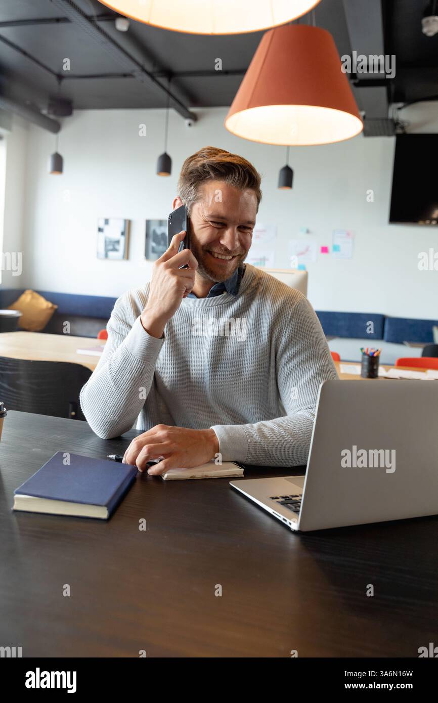 Homme souriant au bureau à l'aide d'un smartphone et d'un ordinateur portable pour la communication d'entreprise Banque D'Images